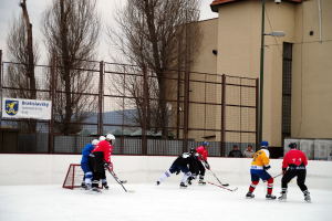 Menschen, die Eis-hockey auf einer Eisbahn mit Gebäuden, Bäumen, einer Straßenlaterne, einem Namensschild und Zäunen im Hintergrund unter einem Himmel spielen.