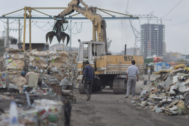 Eine Gruppe von M├Ąnnern, die ├╝ber einen Schotterweg neben einem Haufen M├╝ll gehen, mit einem Bagger im Hintergrund, umgeben von Geb├Ąuden, B├Ąumen, Masten und Dr├Ąhten.