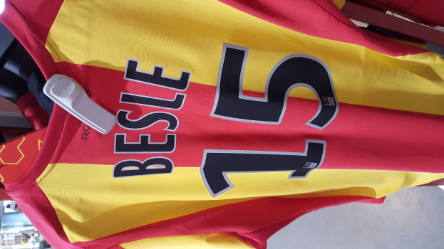 Rotes und gelbes Fußballtrikot mit gedrucktem Text und Nummern an einer Stange hängend, mit anderen Gegenständen und dem Boden im Hintergrund.