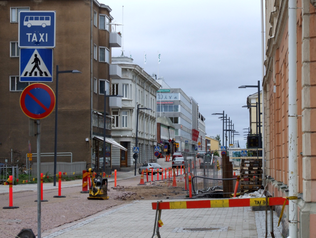 Stadtstraßenszene mit Gebäuden, Fahrzeugen, Verkehrszeichen, Baustelle und Bäumen unter einem bewölkten Himmel.