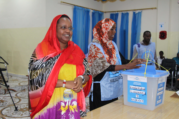 Zwei Frauen stehen nebeneinander an einem Wahltisch mit Papieren und einer Box, während Menschen im Hintergrund sitzen.