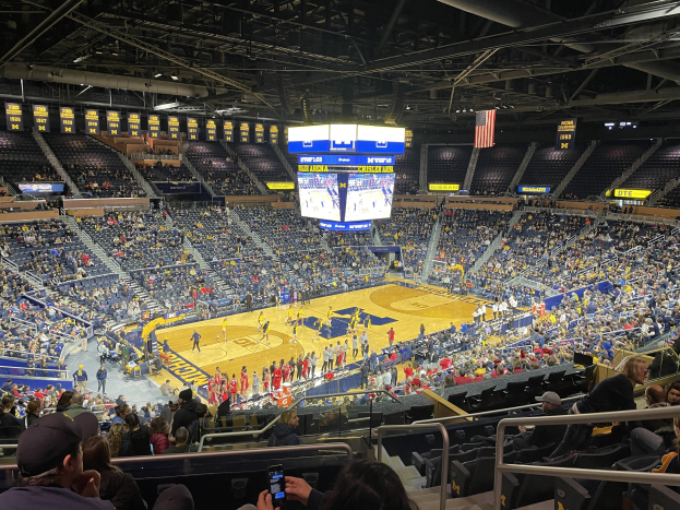 Eine große Arena mit Zuschauern, die ein Basketballspiel verfolgen, mit einer Person, die ein Mobiltelefon hält, Deckenleuchten und Schilder mit der Aufschrift "Michigan Wolverines Basketball".