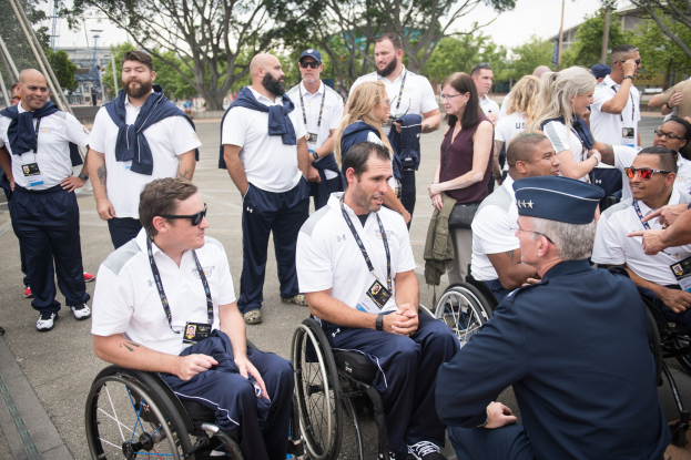 Eine Gruppe von Menschen im Rollstuhl unterhält sich vor einer Kulisse aus Bäumen, Pfählen, Gebäuden und einem klaren blauen Himmel, wobei einige Mützen und Brillen tragen, was darauf hindeutet, dass sie bei den Paralympischen Spielen sind.