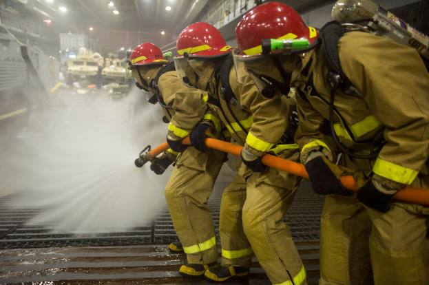 Feuerwehrleute in Helmen und Handschuhen halten Schläuche und spritzen Wasser auf ein Feuerwehrauto, mit Lichtern, Geländern und Tafeln mit Text im Hintergrund.