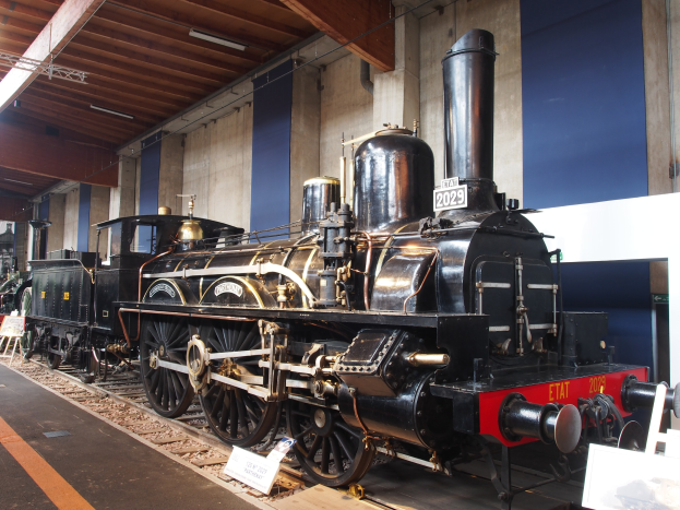 Eine große, alte Dampflokomotive in einem Museums Setting umgeben von Eisenbahnschienen und Informationsschildern, mit beleuchteter Decke und Wand im Hintergrund.