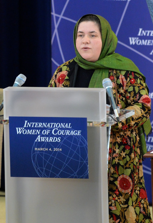 Eine Frau steht an einem Podium mit einem Mikrofon, einer Tafel und einem Tisch zu ihrer Rechten, mit einem Banner mit der Aufschrift "International Women of Courage Awards 2014" im Hintergrund.