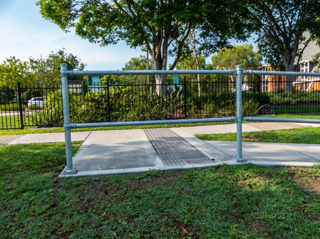 Metallzaun in einer grasbewachsenen Area mit Bäumen und Pflanzen, umgeben von einer Straße mit einem Auto, Gebäude im Hintergrund und einem klaren blauen Himmel.