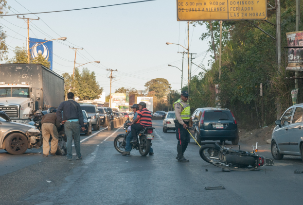 Eine Gruppe von Menschen steht um ein verunglütztes Motorrad auf der Straße mit mehreren Fahrzeugen, darunter ein Lastwagen, und einer Hintergrund von Bäumen, Masten, Lichtern und Schildern unter einem Himmel.