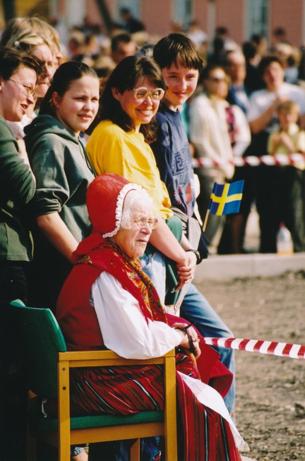 Alte Frau sitzt in einem Stuhl vor einer Menschenmenge, einige tragen Brillen und einer hält eine Fahne, ein Band im Vordergrund und ein leicht unscharfes Gebäude mit Fenstern im Hintergrund.