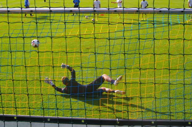 Ein Fußballtorhüter beim Abwehrversuch vor einem Tor, mit einem Netz im Vordergrund und einigen Menschen im Hintergrund, mit verstreuten Fußballen auf dem Boden.