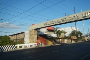 Eine Gruppe von Menschen fährt mit Fahrrädern auf einer Straße in Manila, mit Gebäuden, Bäumen und Strommasten an den Seiten, unter einem bewölkten Himmel und einem "Willkommen in der Stadt Manila"-Bogen.