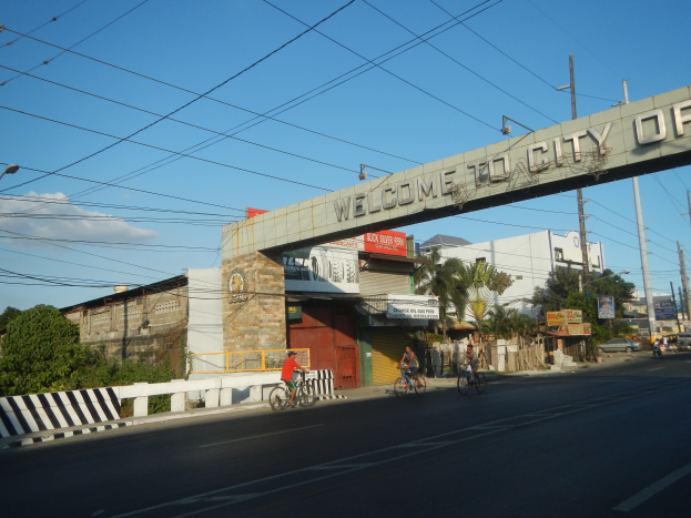 Eine Gruppe von Menschen fährt mit Fahrrädern auf einer Straße in Manila, mit Gebäuden, Bäumen und Strommasten an den Seiten, unter einem bewölkten Himmel und einem "Willkommen in der Stadt Manila"-Bogen.