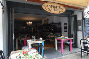 Ein Restaurant im Freien mit Tischen und Stühlen, die mit Tellern, Gläsern und Blumenvasen geschmückt sind, einer Tür, die ins Innere führt, und einer Tafel an der Wand, alles unter einer Decke mit warmem Licht.