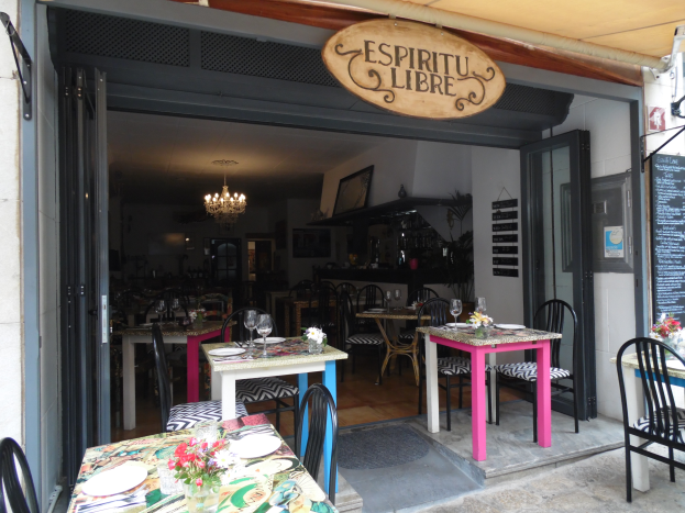 Ein Restaurant im Freien mit Tischen und Stühlen, die mit Tellern, Gläsern und Blumenvasen geschmückt sind, einer Tür, die ins Innere führt, und einer Tafel an der Wand, alles unter einer Decke mit warmem Licht.