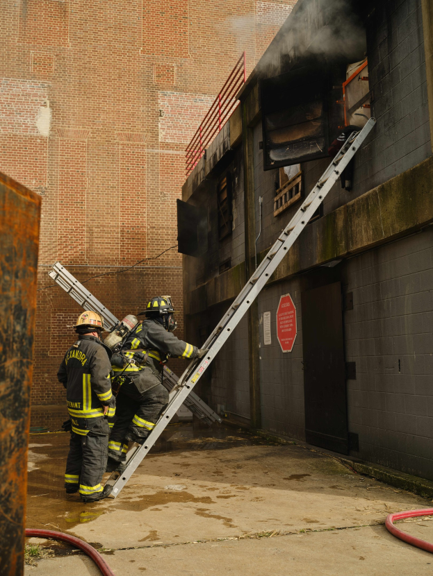 Feuerwehrleute in Helmen klettern an einer Leiter zu einem rauchgefüllten Gebäude, mit einem Metallobjekt links und Rohren an der Basis.