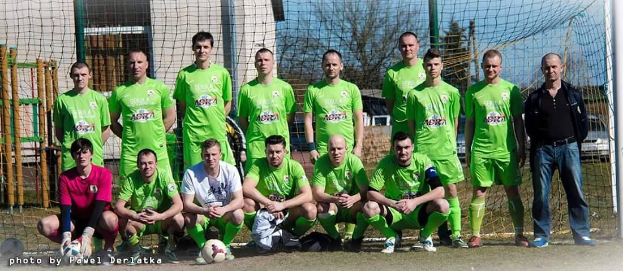 Gruppe von Männern in grünnen T-Shirts auf einem Fussballfeld mit einem Ball und einem Torpfosten, Bäume und Gebäude im Hintergrund.