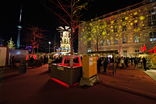 Ein lebendiger Weihnachtsmarkt in Berlin, Deutschland, mit Menschen um beleuchtete Buden, Bäume, Gebäude, Laternenmasten und einen Turm unter einem dunklen Himmel.