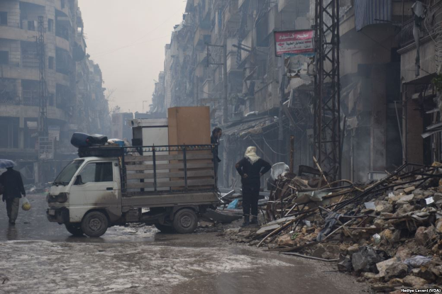 Ein Lastwagen fährt auf einer Straße neben einem Schutthaufen, mit Menschen auf der Straße, einer hält einen Schirm, und Gebäuden, Polen, Brettern mit Text im Hintergrund unter einem klaren Himmel.