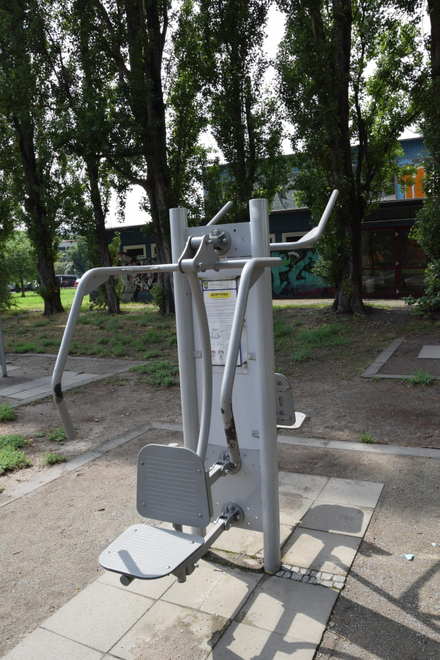 Ein Park mit Fitnessgeräten in der Mitte, umgeben von Bäumen und Gebäuden unter einem klaren blauen Himmel.