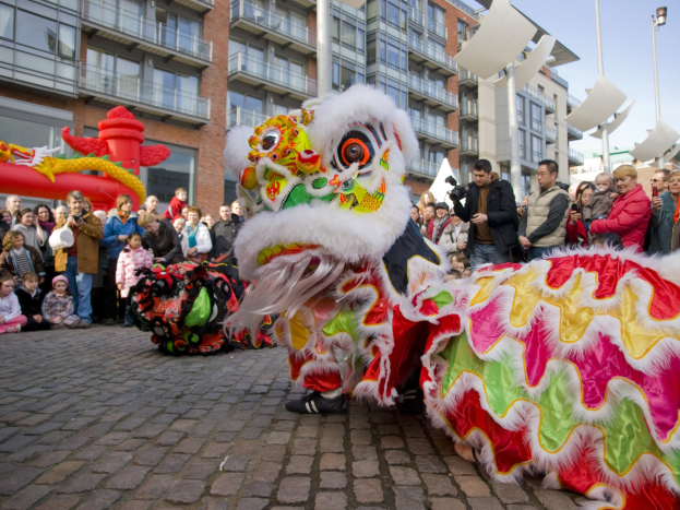Ein lebendiges chinesisches Neujahrsfest in Amsterdam mit einer Löwen-Tanz-Show vor einem Publikum, das Kameras hält, vor Gebäuden und Laternenmasten unter einem klaren blauen Himmel.
