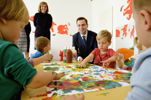 Kinder beim Malen um einen Tisch mit Farbe an den Händen, beaufsichtigt von einer lächelnden Frau, mit Kunstwerken an der Wand.