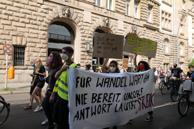 Eine Gruppe von Menschen marschiert auf einer Stadtstraße bei einer Demonstration, einige halten Schilder und andere fahren Fahrräder, mit einem historischen Gebäude, Bäumen und einer Straßenlaterne im Hintergrund.