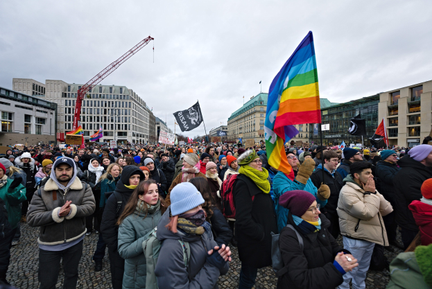 Eine große Gruppe von Menschen steht vor einem Gebäude und hält Fahnen und Schilder mit der Aufschrift 'Lgbtq+-Rechte-Demo in Berlin' in den Händen, einige tragen Mützen und Taschen, während im Hintergrund Gebäude, ein Kran und Wolken zu sehen sind.