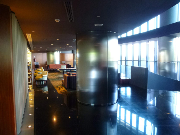 Eine geräumige First-Class-Lounge am Hong Kong International Airport mit großen Fenstern, hohen Decken mit elektrischen Leuchten, Sitzmöglichkeiten einschließlich Stühlen und Sofas, Tischen, einem Teppichboden und einer mittigen Säule.