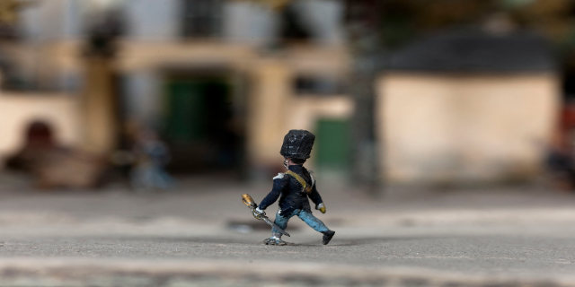 Eine Spielzeugsoldat mit einer Pistole in der Hand geht eine Straße entlang mit unscharfen Gebäuden im Hintergrund.