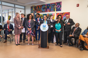 Gruppe von Menschen vor einem Rednerpult mit einem Mikrofon und einer Tafel mit der Aufschrift "Sancho-Stadtrat tritt dem Stadtrat bei", mit sitzenden und stehenden Teilnehmern, Flaggen, einem Feuerlöscher und Deckenleuchten an der Rückwand.