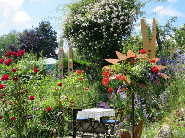 Ein Garten auf der Chelsea Flower Show mit bunten Blumen und Pflanzen, einem Tisch mit einer Decke und Stühlen in der Mitte und Bäumen im Hintergrund unter einem bewölkten Himmel.