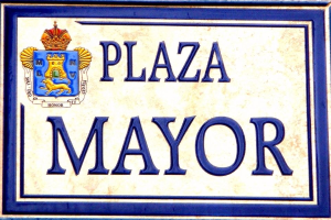 Rechtwinkliges weißes Schild mit schwarzer "Plaza Mayor"-Beschriftung, blauem Rahmen und einem gekrönten Wappen-Logo an einer Gebäudefassade.