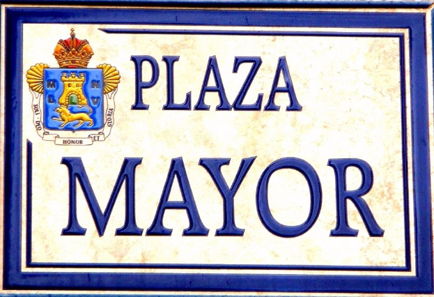 Rechtwinkliges weißes Schild mit schwarzer "Plaza Mayor"-Beschriftung, blauem Rahmen und einem gekrönten Wappen-Logo an einer Gebäudefassade.