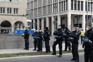 Eine Gruppe von Polizisten in schwarzen Uniformen und Masken steht vor einem Gebäude mit Glasfenstern und Säulen in Berlin, Deutschland, einige halten Helme, mit Laternen, Verkehrsampeln, Schildern mit Text, einer Statue, anderen Menschen und grasbewachsenem Boden im Hintergrund.
