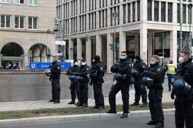 Eine Gruppe von Polizisten in schwarzen Uniformen und Masken steht vor einem Gebäude mit Glasfenstern und Säulen in Berlin, Deutschland, einige halten Helme, mit Laternen, Verkehrsampeln, Schildern mit Text, einer Statue, anderen Menschen und grasbewachsenem Boden im Hintergrund.