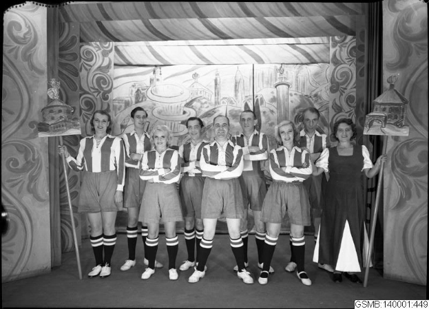 Schwarzes und weißes Foto einer Frauenfußballmannschaft der 1940er Jahre auf einer Bühne, die strahlt und Stöcke hält, mit einer bemalten Wand und Text unten.