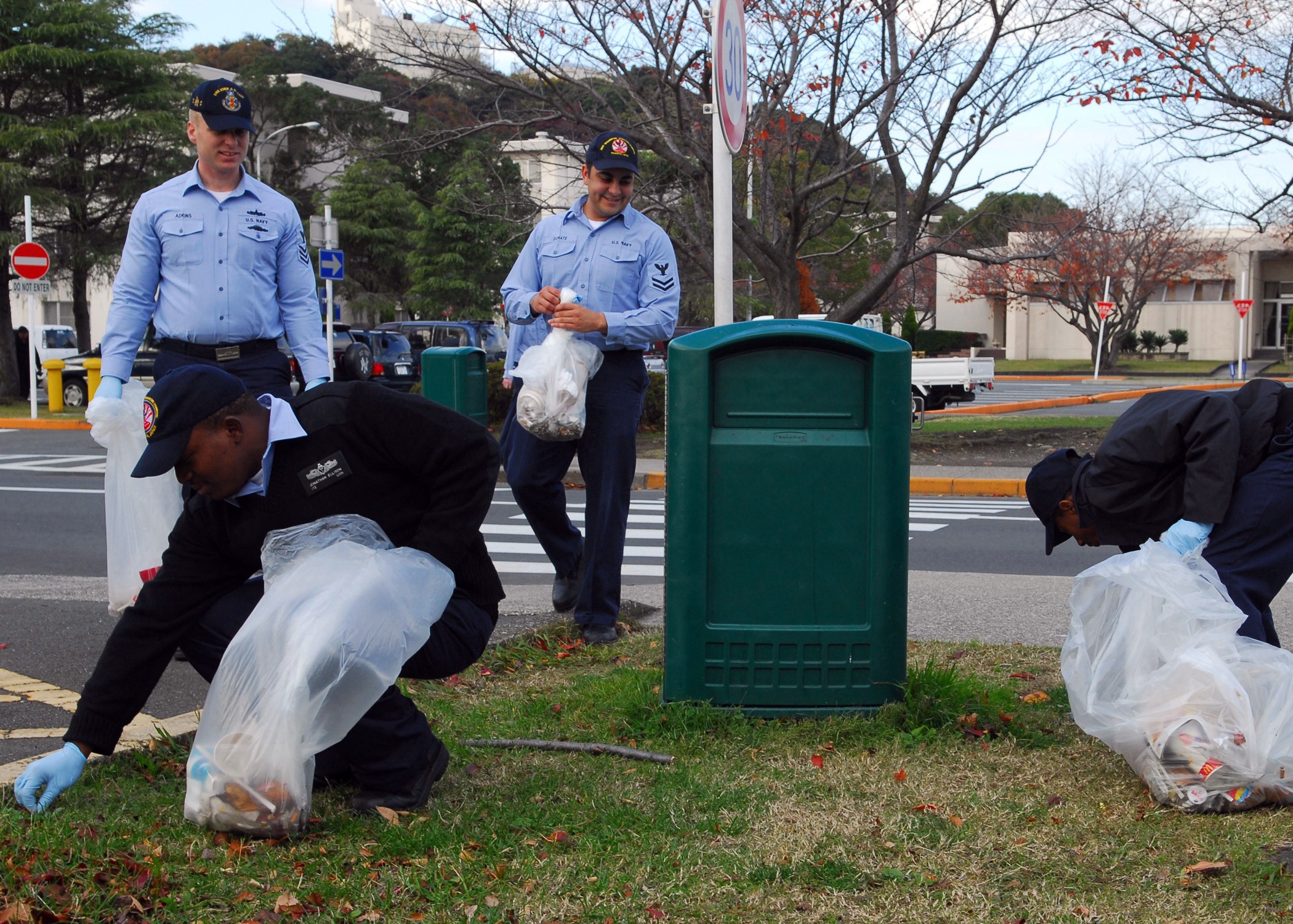 Drei Polizeibeamte in Uniform sammeln Müll am Straßenrand mit Bäumen, Gebäuden und Fahrzeugen im Hintergrund.