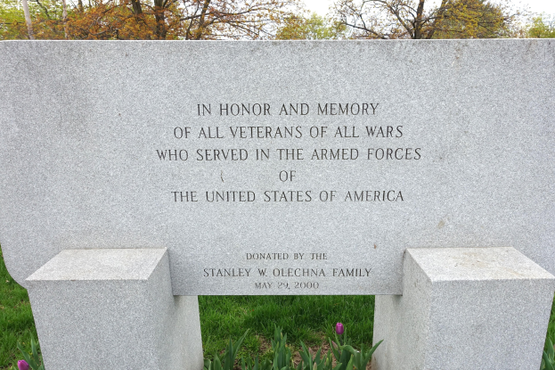 Denkmalstein mit einer Inschrift für US-Streitkräfte-Veteranen, umgeben von Pflanzen, Blumen, Gras, Bäumen und einem bewölkten Himmel.