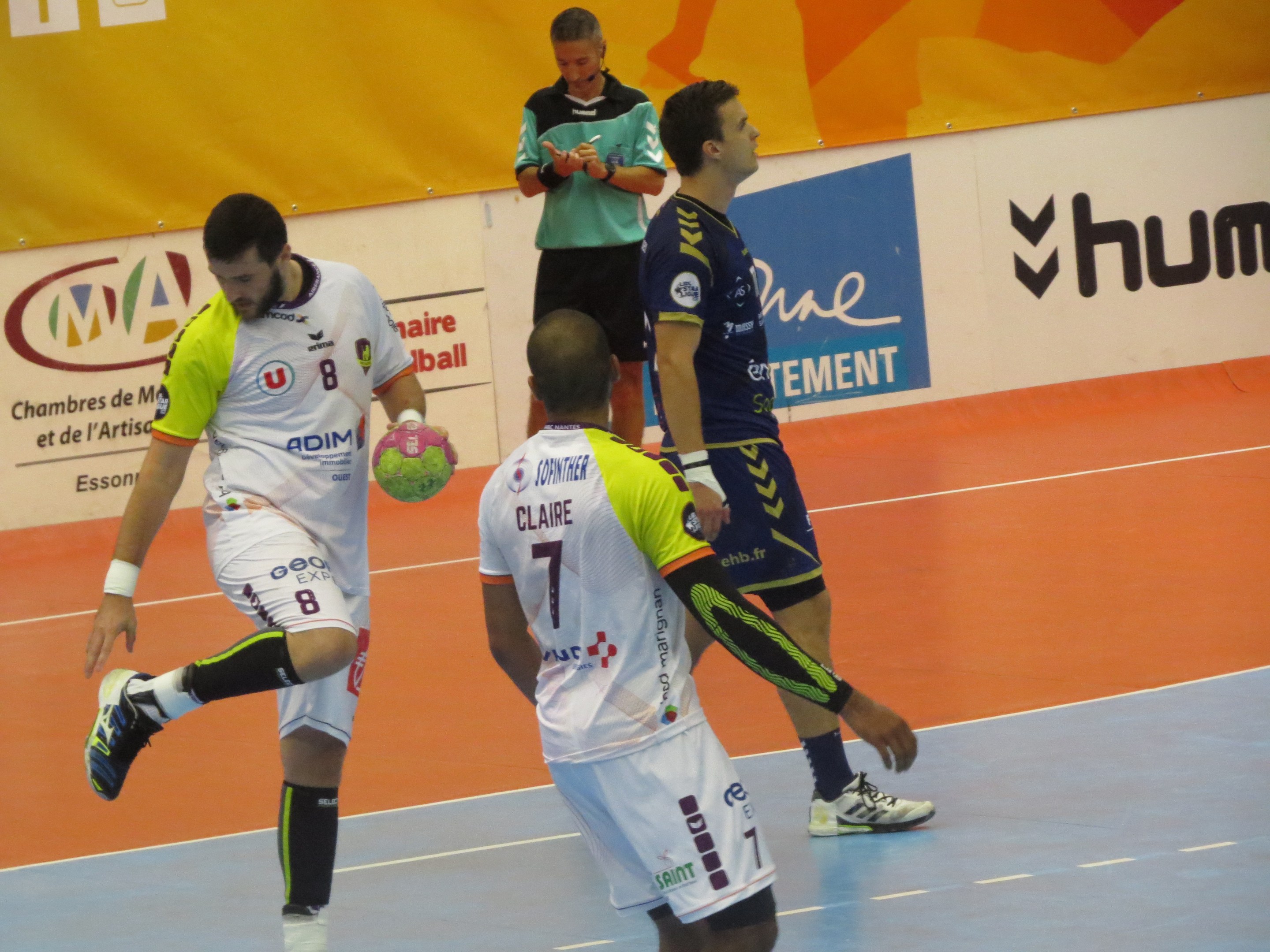 Mannergruppe spielt Handball auf einem Platz, einer hält den Ball, mit Bannern im Hintergrund.